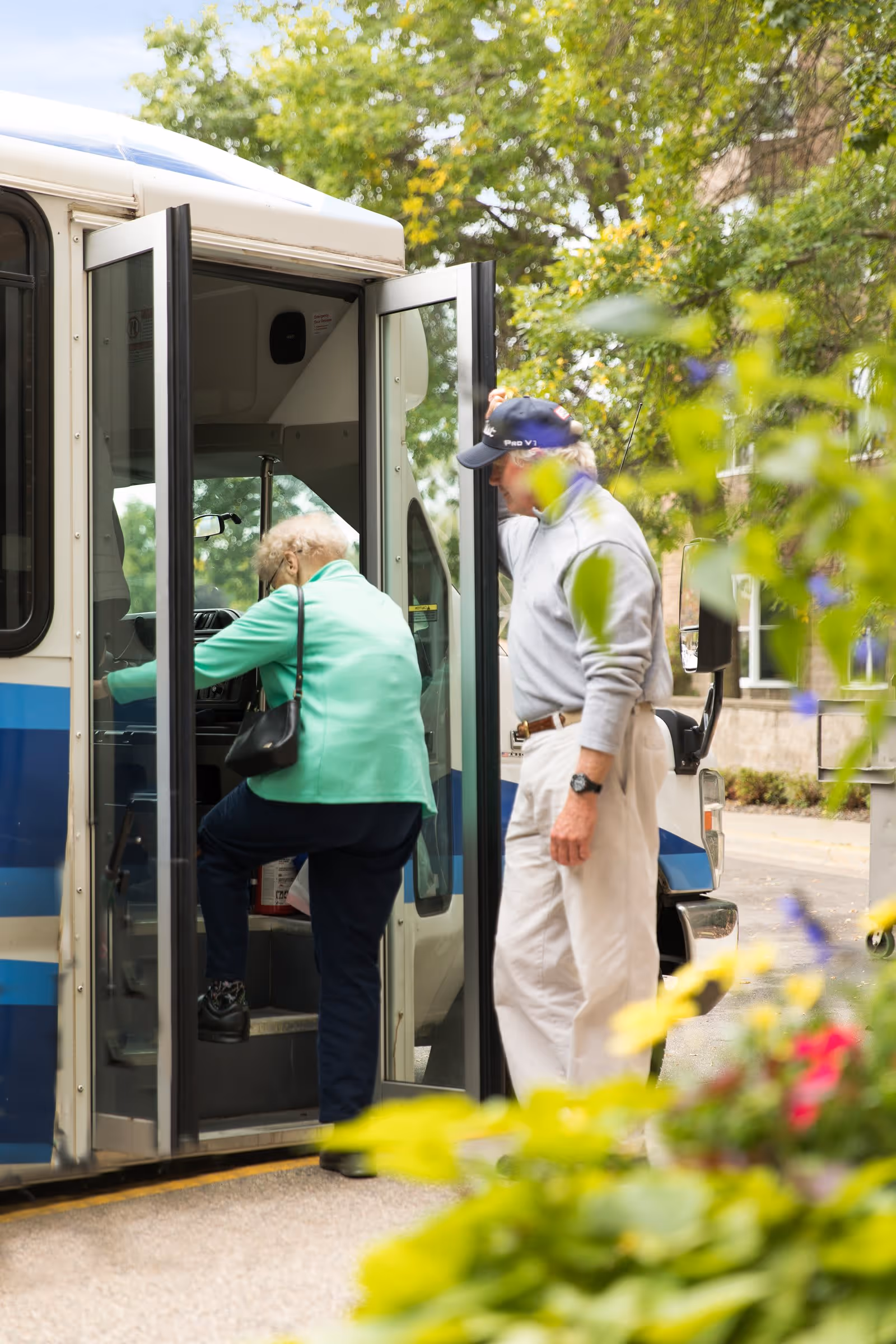 An elderly woman wearing a green jacket and carrying a black purse is stepping onto a bus, while an elderly man in a light gray sweater and white pants stands nearby holding the bus door open. The scene is outdoors with greenery and flowers in the foreground and trees in the background.