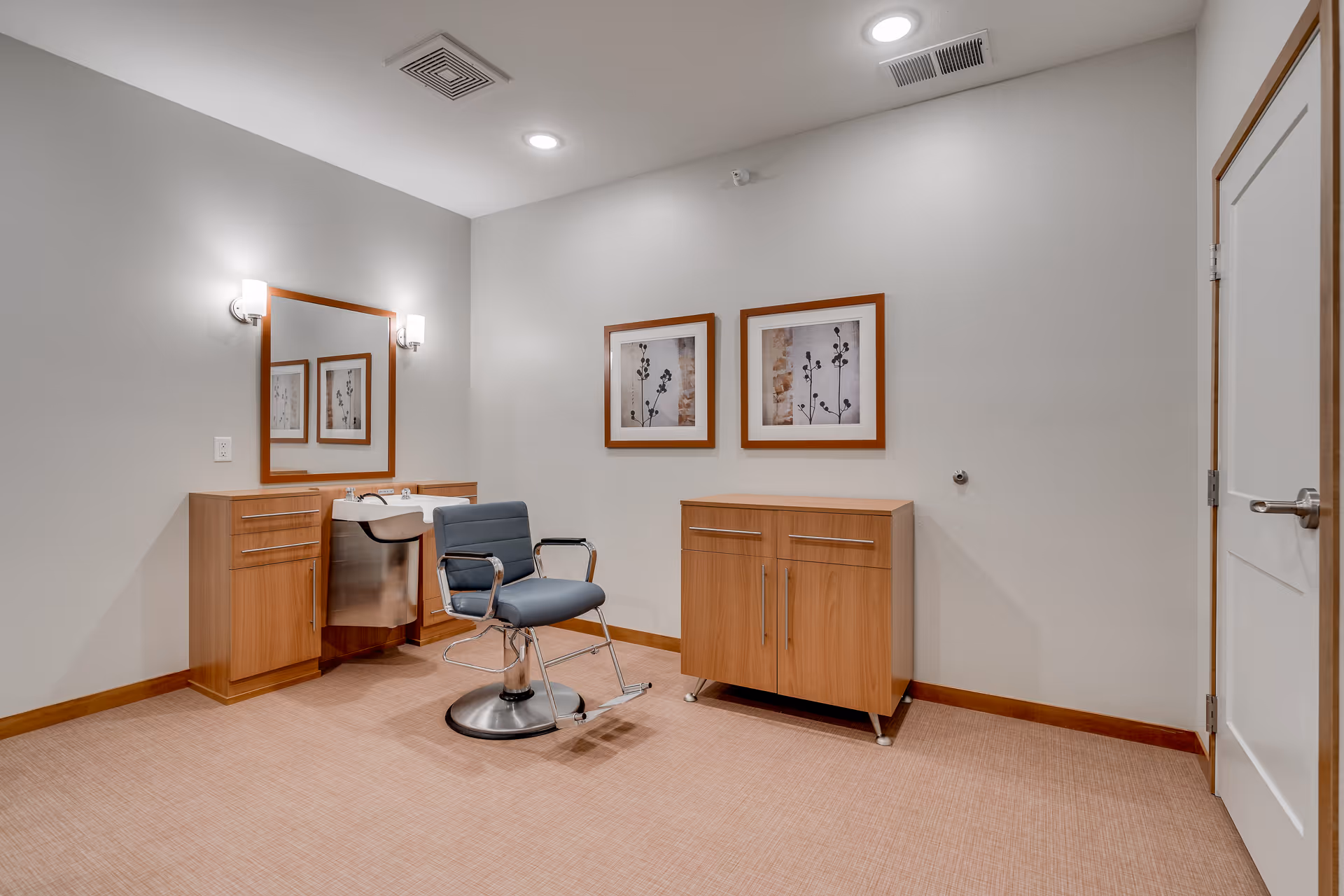 A clean and modern salon area in a senior living facility featuring a single salon chair with armrests and footrest, a large mirror mounted on the wall above a wooden cabinet with a built-in sink, two framed botanical prints on the wall, and a closed white door on the right side.