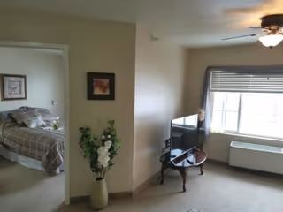Interior view of a senior living facility room showing a living area with a TV on a small wooden table near a window with blinds. To the left, there is an open doorway leading to a bedroom with a bed covered in a plaid bedspread. A tall vase with white flowers and green leaves is placed against the wall near the doorway. The room has beige walls and carpeted floor.