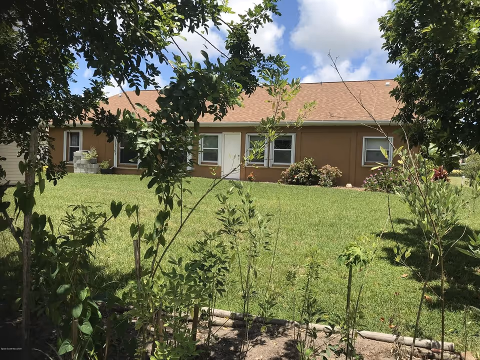 Single-story brown house with a front lawn and shrubs seen through foreground trees and plants.
