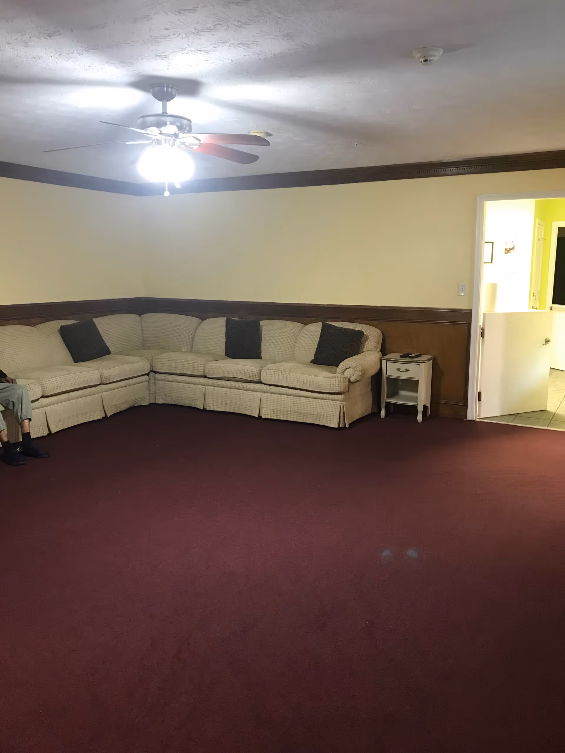 A living room with a beige sectional sofa adorned with three dark cushions, a small white side table next to the sofa, maroon carpet flooring, cream-colored walls with dark wood paneling halfway up, and a ceiling fan with lights turned on. An open door reveals a glimpse of a hallway with yellow walls and tiled floor.