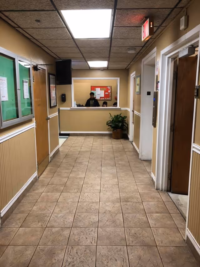 Interior hallway of a facility with beige walls and tiled floor leading to a reception window where two people are visible. There are doors on both sides of the hallway, a potted plant near the reception, and an exit sign above a door on the right.