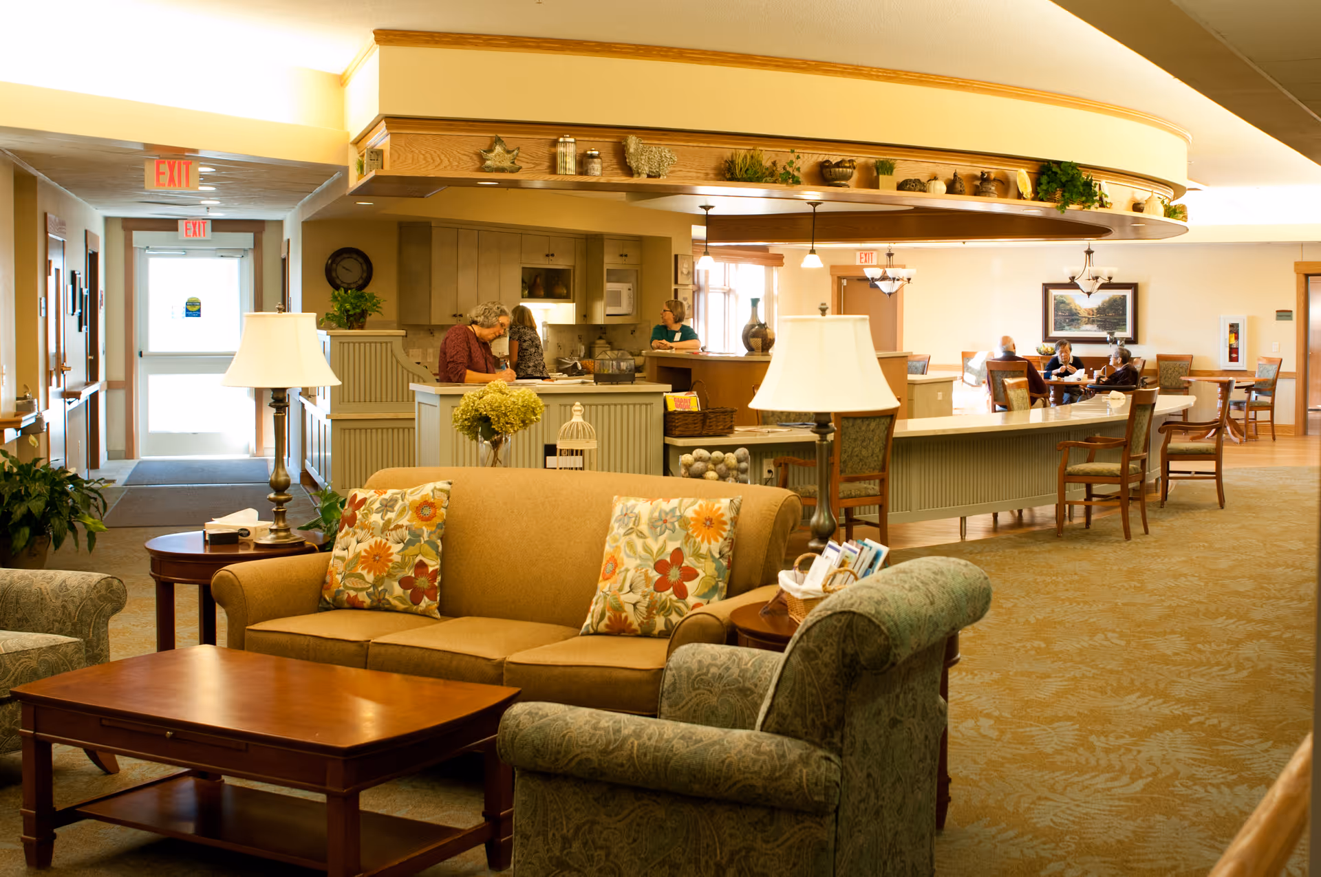 Warmly lit senior living common area with sofas and armchairs in the foreground and a reception/dining counter with residents and staff in the background.