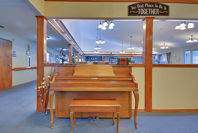 Interior view of a senior living facility showing a wooden upright piano with a matching bench placed against a half wall with wooden columns. Above the piano, a sign reads 'The Best Place To Be Is TOGETHER'. The background reveals a spacious common area with multiple ceiling lights and large windows.