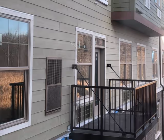 Exterior of a residential building showing siding, multiple windows, a wall-mounted vent/heater, and a small metal balcony.