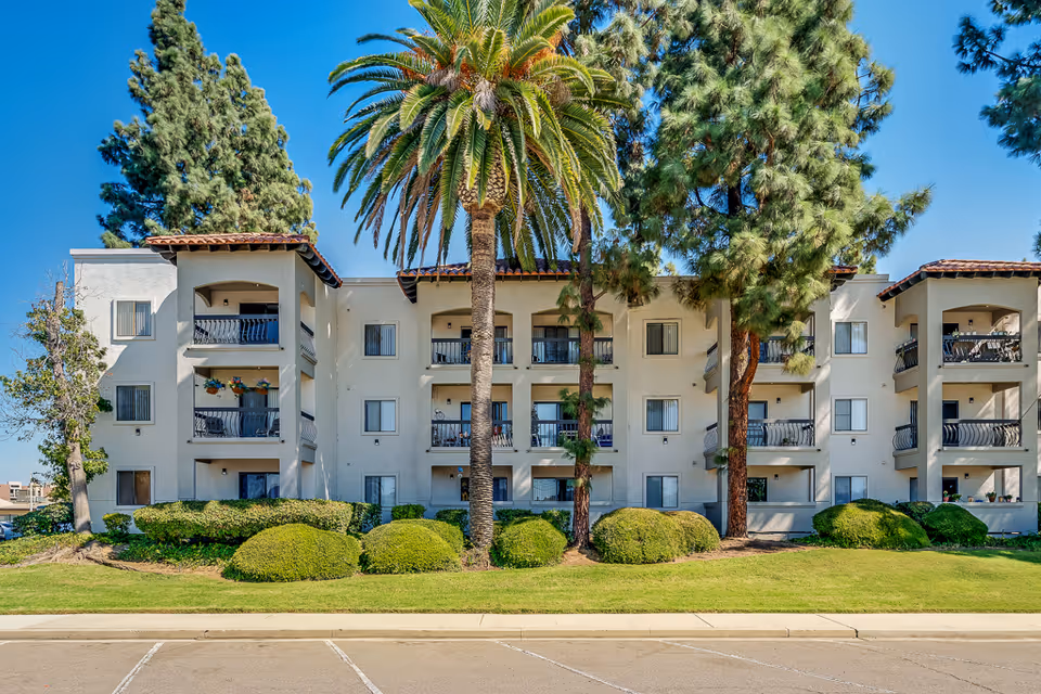 White three-story Mediterranean-style apartment building with balconies, palm trees, and landscaped shrubs in front.
