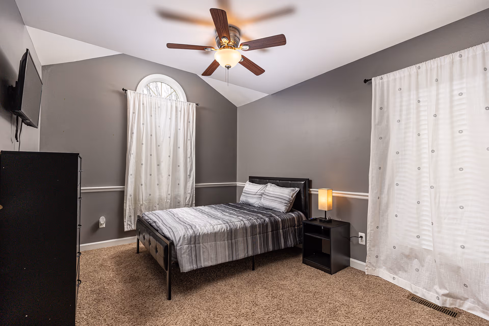 A bedroom with gray walls and beige carpet featuring a bed with a striped gray and white comforter and two pillows. There is a black nightstand with a lamp on the right side of the bed, a black dresser on the left side, and a wall-mounted TV above the dresser. The room has two windows covered with white curtains, one of which is arched at the top. A ceiling fan with wooden blades and a light fixture is mounted on the ceiling.