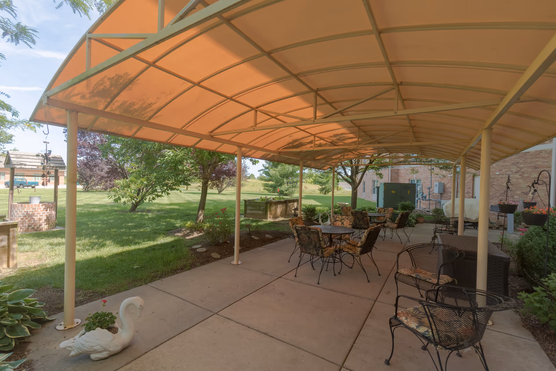 Outdoor covered patio area with several metal chairs and tables with floral cushions. The patio is adjacent to a grassy lawn with trees and garden beds. A decorative white swan planter is visible on the concrete floor near the edge of the patio.