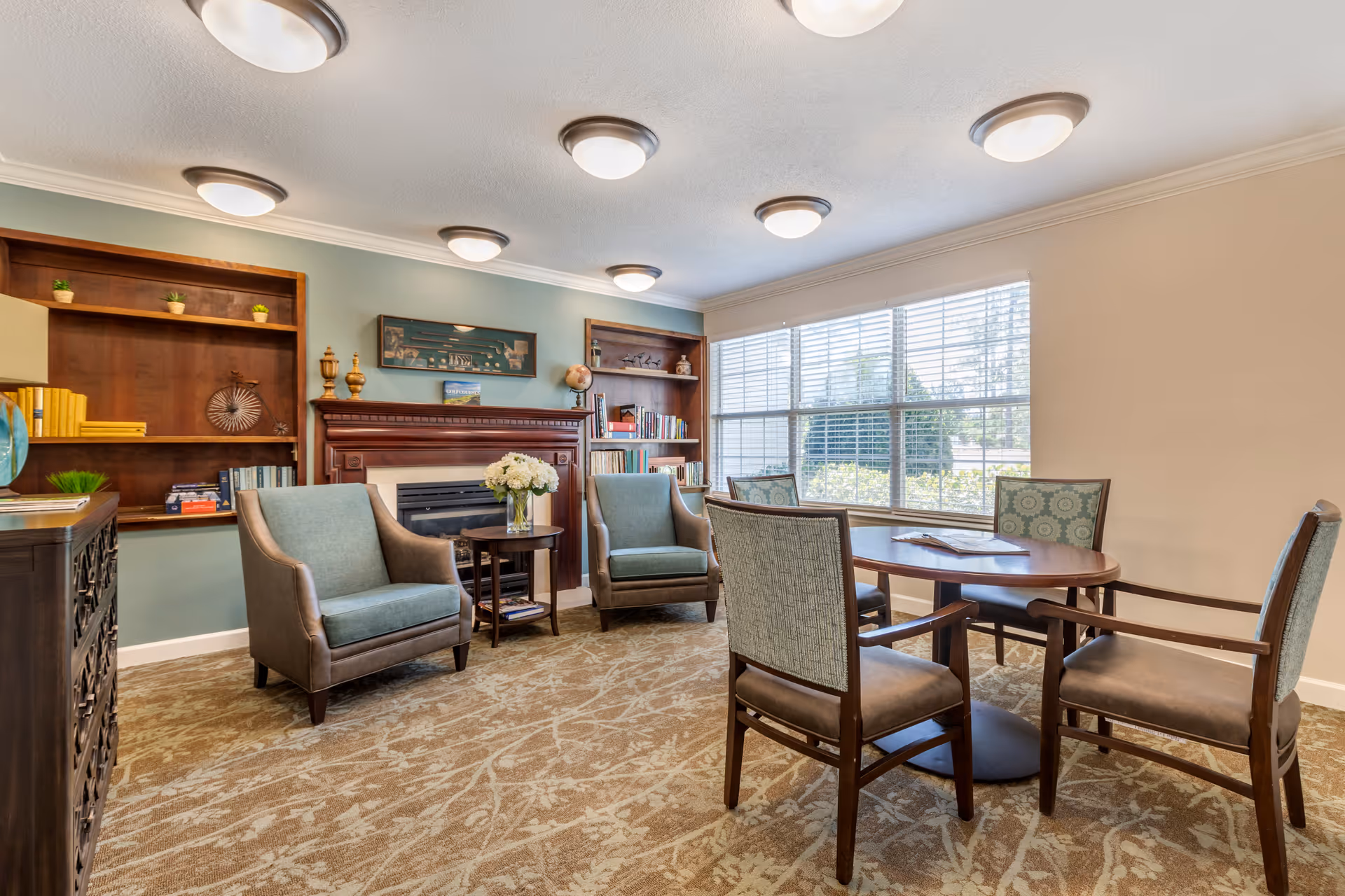 A bright common room with upholstered armchairs and a round table in front of a fireplace and built-in bookshelves beside a large window.