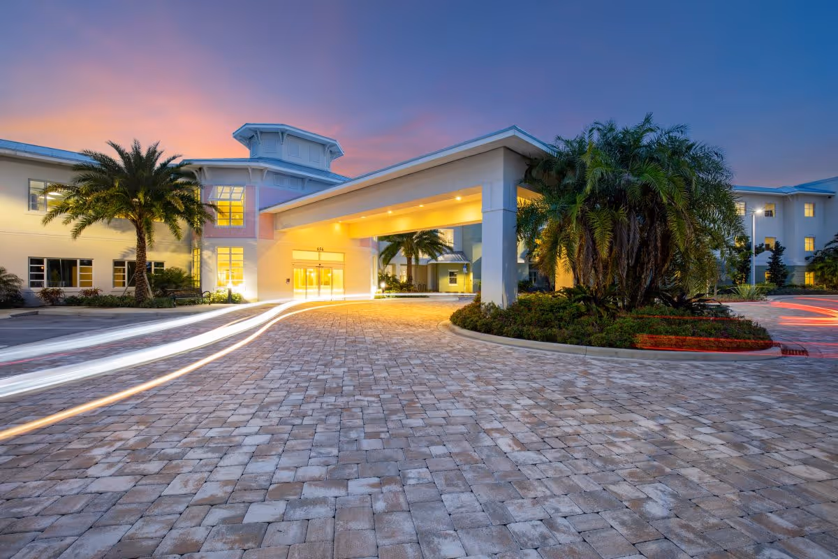 Front entrance of a senior living building with a covered porte-cochère, palm trees, and illuminated windows at dusk.