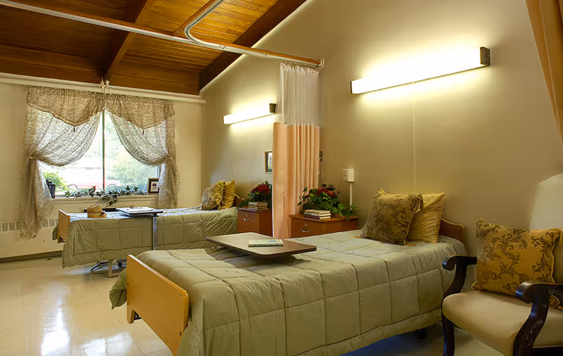 A cozy room with two single beds covered in light green quilts, each with decorative pillows. There is a small wooden nightstand between the beds with books and a plant on top. A window with sheer curtains lets in natural light, and a beige armchair with patterned cushions is placed near one of the beds. The room has warm lighting from wall-mounted fixtures and a wooden ceiling.