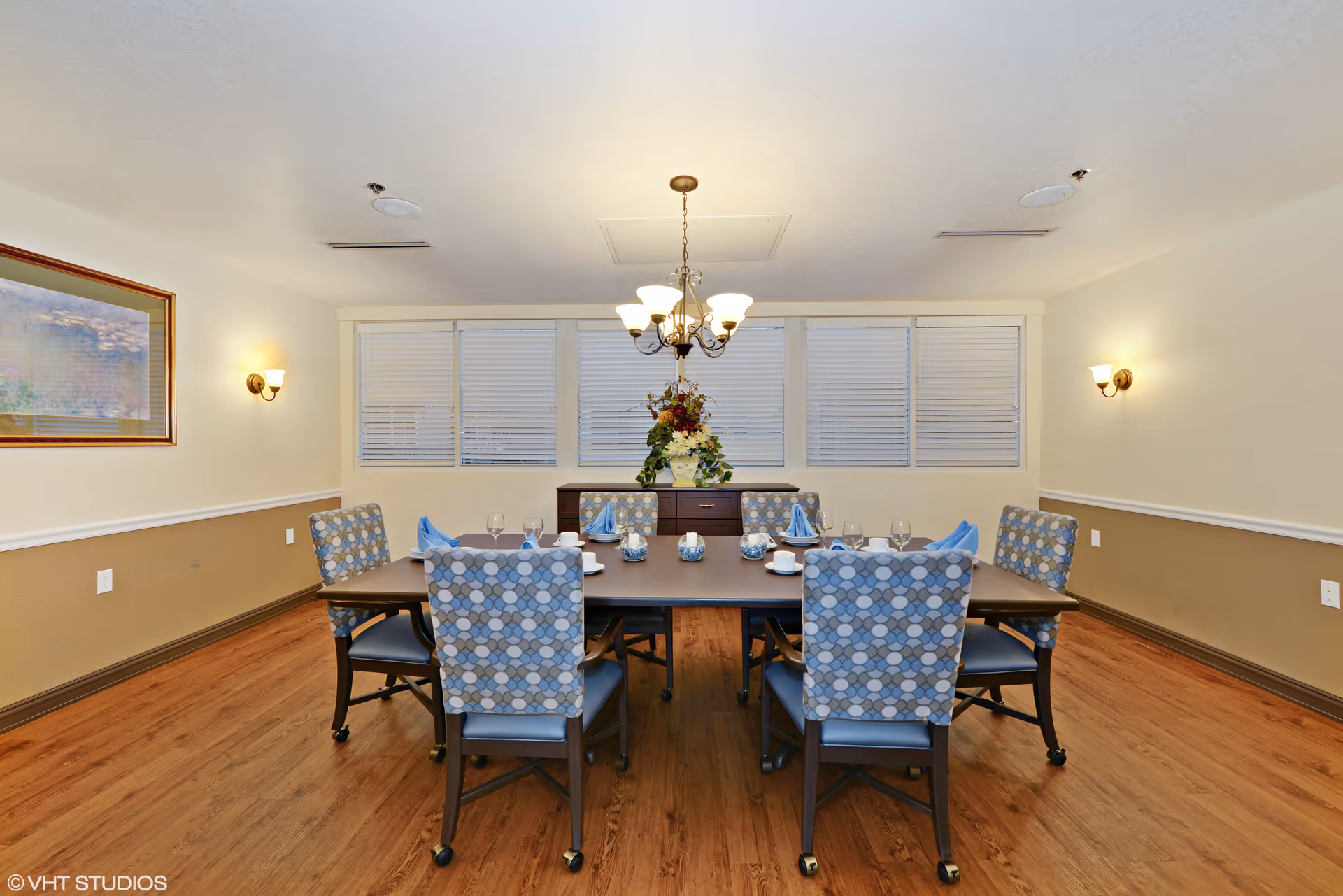 A dining room with a rectangular table set for six, featuring patterned chairs with wheels, blue napkins, glassware, and white cups. The room has wooden flooring, beige and white walls, a chandelier hanging from the ceiling, wall sconces, a framed picture on the left wall, and a sideboard with a floral arrangement in front of a row of windows with blinds.