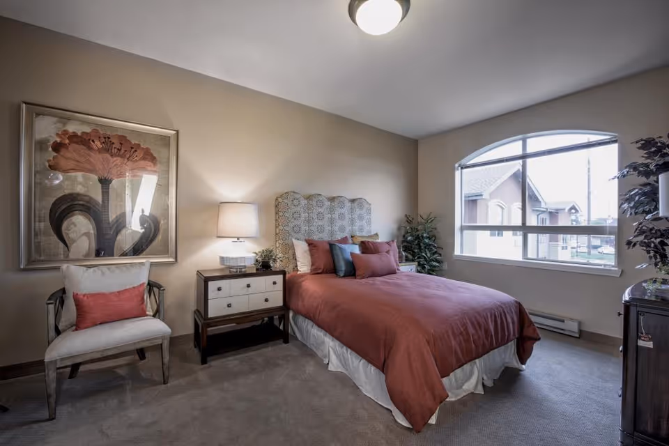 A cozy bedroom with a large window letting in natural light. The room features a bed with a patterned headboard, red bedding, and multiple pillows. Next to the bed is a nightstand with a lamp and small plant. A cushioned chair with a red pillow sits beside the nightstand. A large framed floral artwork hangs on the wall above the chair. There is a potted plant near the window and a small dark wooden cabinet on the right side of the room.