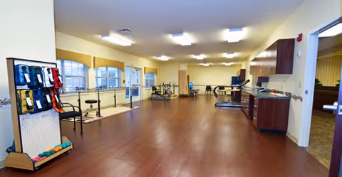 A spacious rehabilitation or therapy room with wooden flooring, exercise equipment including a treadmill and stationary bike, parallel bars for walking support, and a rack holding various therapy bands and balls. The room has large windows allowing natural light and overhead fluorescent lighting. There is a countertop with cabinets on the right side and an open doorway leading to another room.