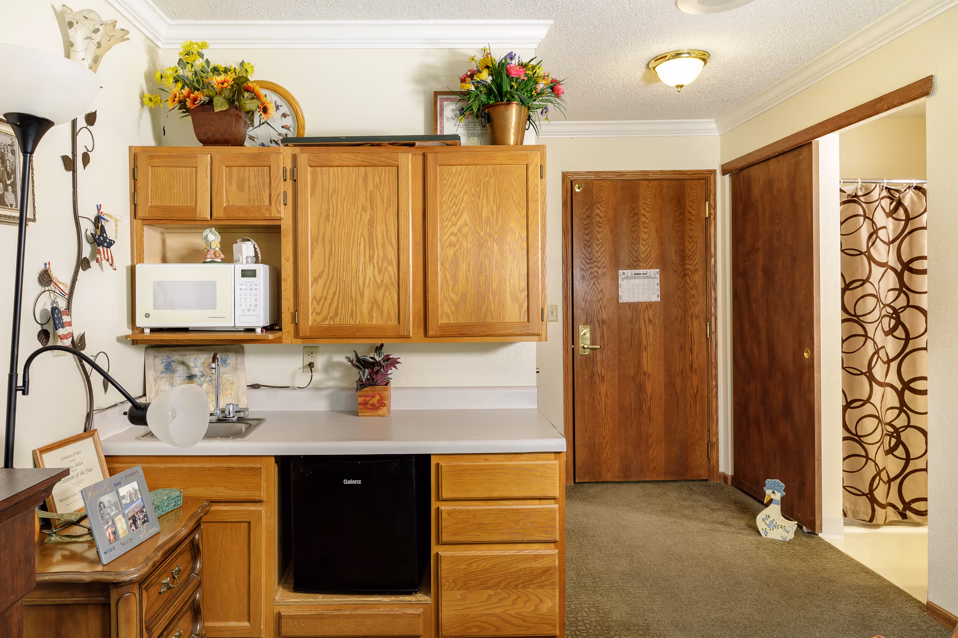 Interior view of a small kitchenette area with wooden cabinets, a white microwave, a small black refrigerator, and a countertop with a small sink. There are decorative flowers on top of the cabinets and a lamp on a wooden dresser to the left. A wooden door and a sliding door leading to a bathroom with a shower curtain are visible in the background.
