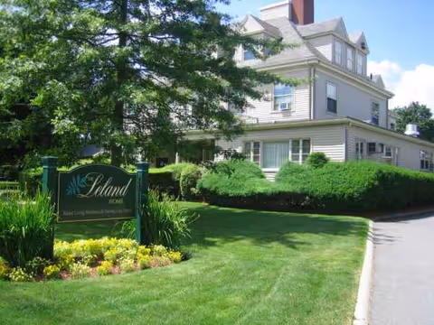 Two-story light-colored senior living building with manicured lawn and a sign reading Leland House.