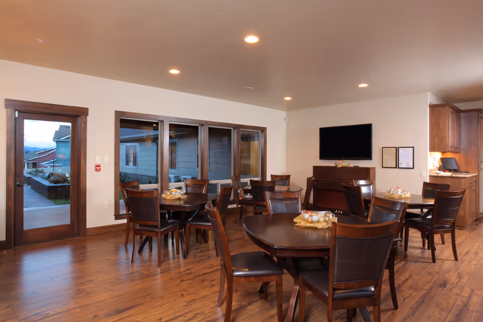 Interior view of a dining room with several round wooden tables and chairs arranged neatly. There is a piano against the far wall beneath a mounted flat-screen TV. Large windows and a glass door provide a view of the outdoor area. The room has wooden flooring and recessed ceiling lights.