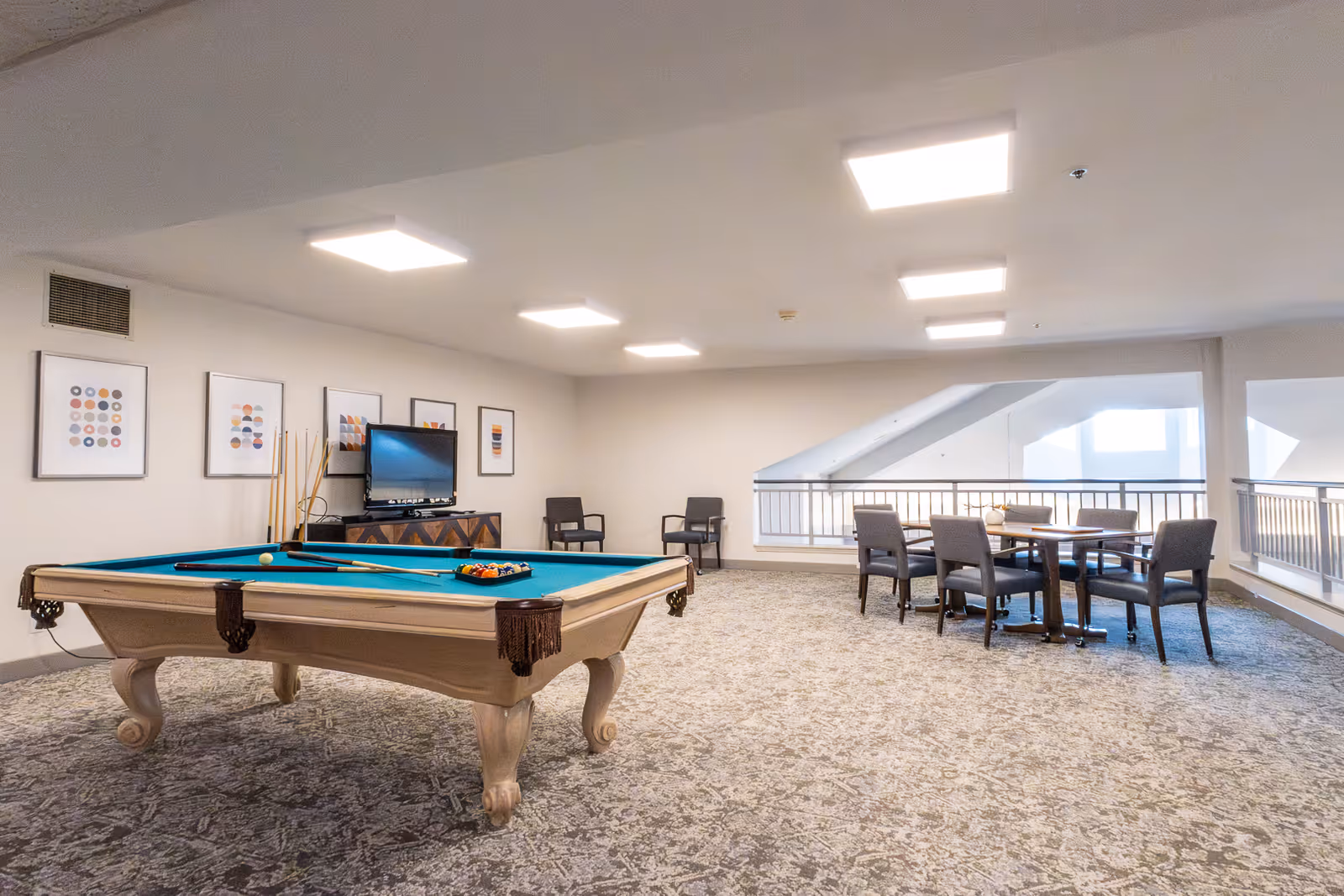 A spacious recreational room with a pool table in the center, a TV on a stand against the wall, several framed abstract art pieces hanging above the TV, and a seating area with a table and chairs near a railing overlooking a lower level.