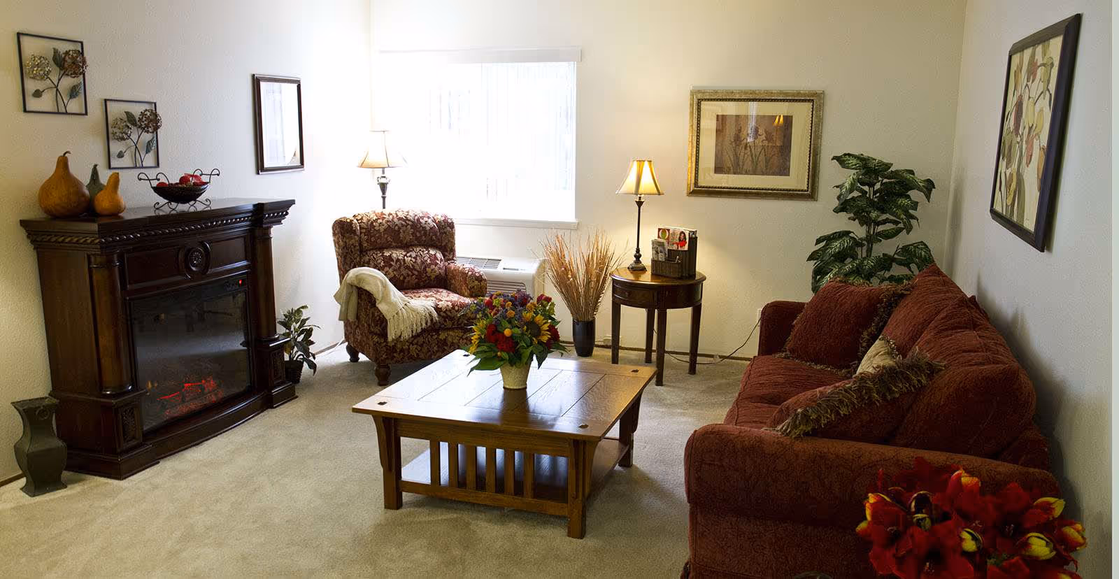 A cozy living room with a patterned armchair and a red sofa with matching pillows. A wooden coffee table with a flower arrangement sits in the center. There is a dark wooden electric fireplace on the left side with decorative gourds and a fruit bowl on top. Two table lamps illuminate the room, one on a small round wooden side table and the other near the window with vertical blinds. The walls are decorated with framed artwork and there is a green potted plant in the corner.
