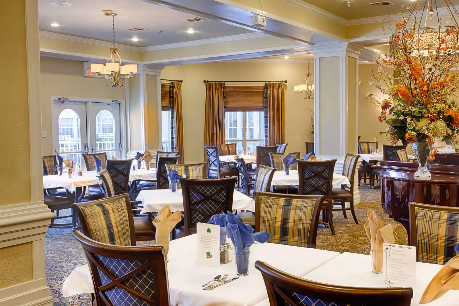 Bright dining room with multiple tables set with white linens and napkins, upholstered chairs, and a large floral centerpiece.