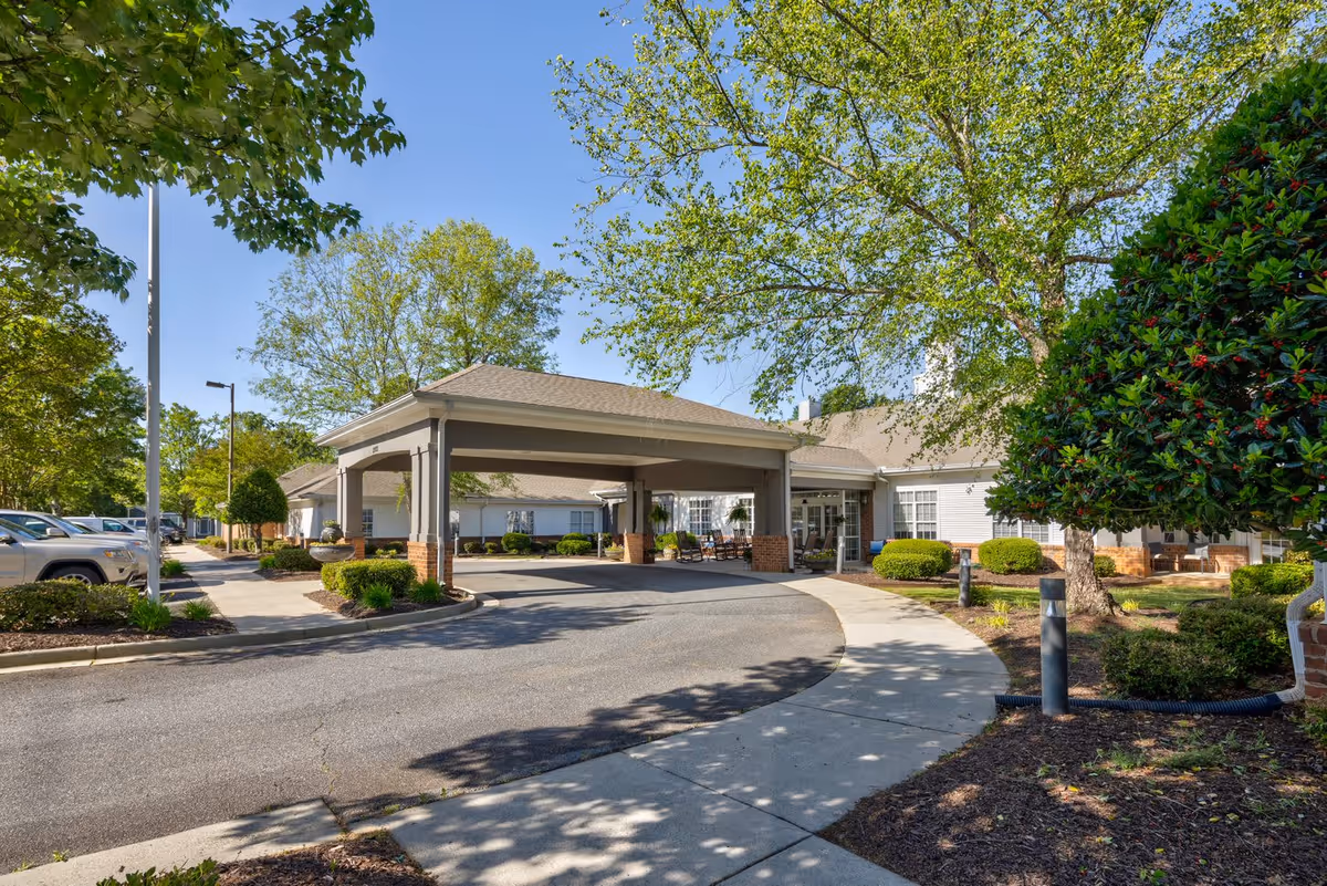 Exterior view of TerraBella Marchbanks senior living facility showing the main entrance with a covered drop-off area, surrounded by trees, shrubs, and a parking area with cars under a clear blue sky.