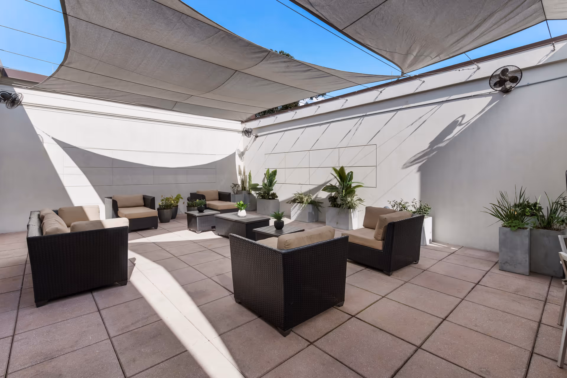 Outdoor patio area with cushioned wicker chairs and sofas arranged around coffee tables, shaded by large fabric canopies overhead. Several potted plants line the white walls, and two wall-mounted fans are visible.