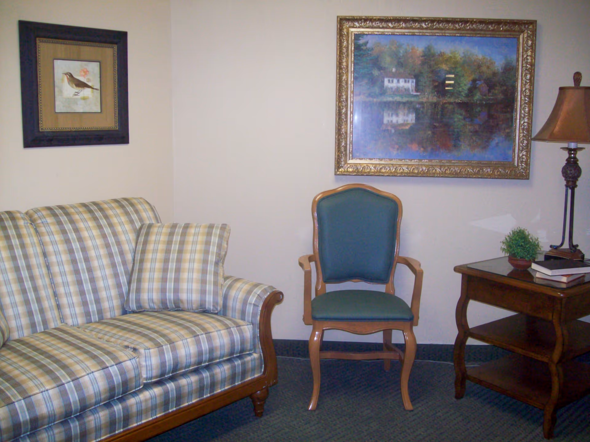 A cozy sitting area with a plaid upholstered sofa, a wooden framed green cushioned chair, a wooden side table with books and a small plant, a floor lamp, and two framed paintings on the wall.