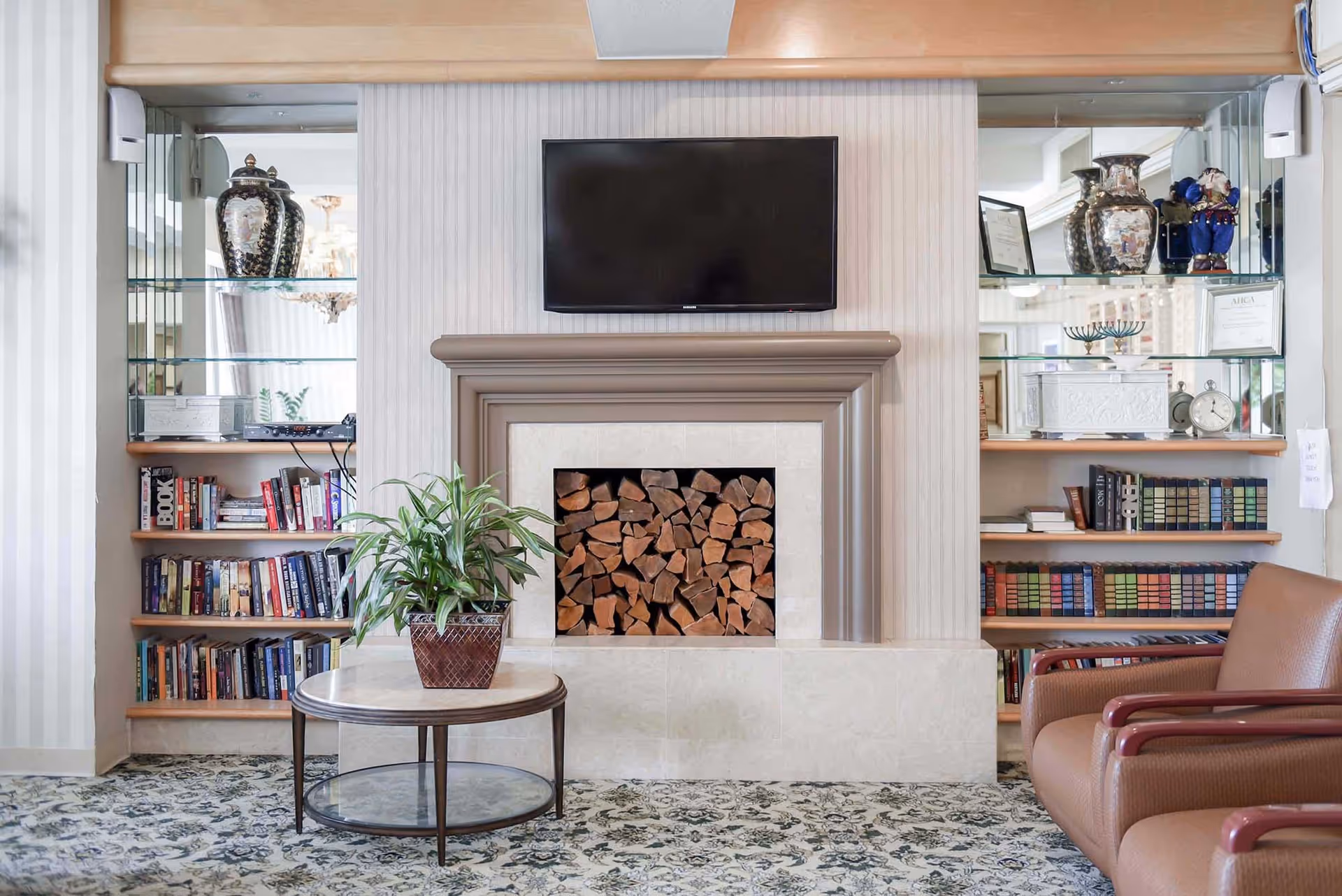 A cozy living room area featuring a fireplace filled with stacked firewood, a flat-screen TV mounted above the fireplace, built-in shelves on either side with books, decorative vases, framed certificates, and a small clock. In front of the fireplace is a round coffee table with a potted plant, and to the right, there are two brown armchairs with wooden armrests. The room has patterned carpet flooring and light-colored striped wallpaper.