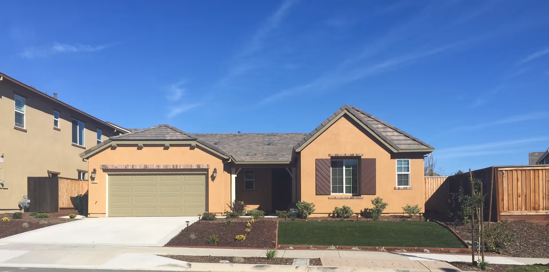 Single-story house with a beige exterior, a two-car garage with a green door, a small front lawn with shrubs, and a clear blue sky above.