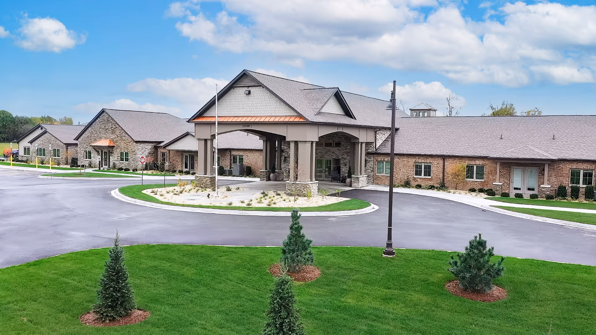 Front exterior view of Cedarhurst Senior Living of Frankfort showing a large building with a covered entrance, brick and stone facade, surrounded by a paved driveway and well-maintained green lawn with small trees under a partly cloudy sky.