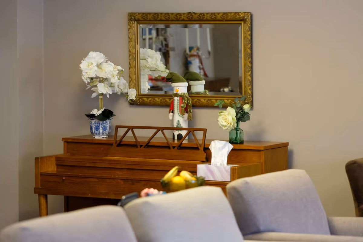 A wooden piano with a decorative music stand, topped with a white orchid in a blue and white pot, a snowman figurine wearing a green hat and scarf, a white rose in a green vase, and a box of tissues. Above the piano is a gold-framed mirror reflecting part of the room. In the foreground, there are beige upholstered chairs.