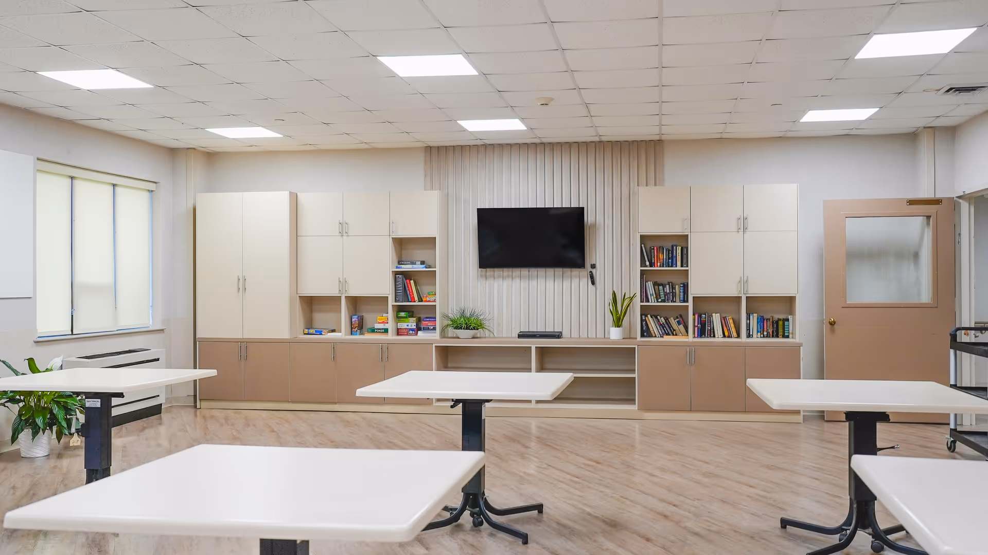 Bright multipurpose activity room with white tables, built-in cabinets, bookshelves and a wall-mounted TV.