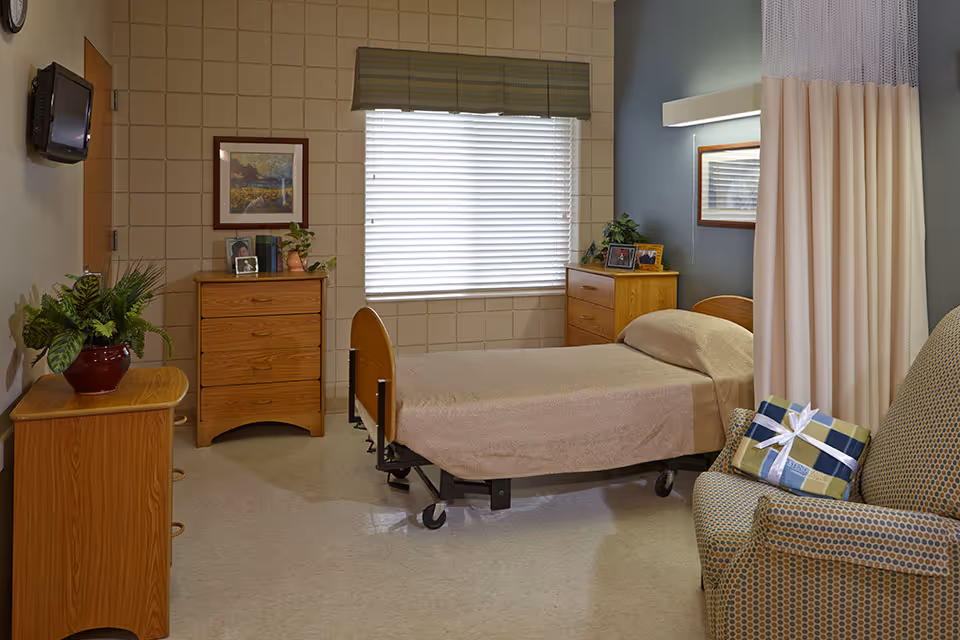 A senior living facility bedroom with a single hospital-style bed covered with a beige blanket, two wooden dressers with framed photos and plants on top, a window with blinds and a valance, a patterned armchair with a wrapped gift on it, a wall-mounted TV, and a privacy curtain partially drawn.