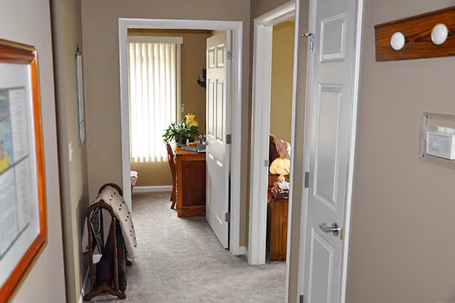 View down a carpeted hallway in a residential care facility with beige walls and white doors. On the left side, there is a coat rack with a jacket hanging on it and a framed picture on the wall. At the end of the hallway, there is a room with a wooden desk, a chair, and a window with vertical blinds. On the right side, two open doors reveal parts of rooms with furniture including a lamp and a side table.