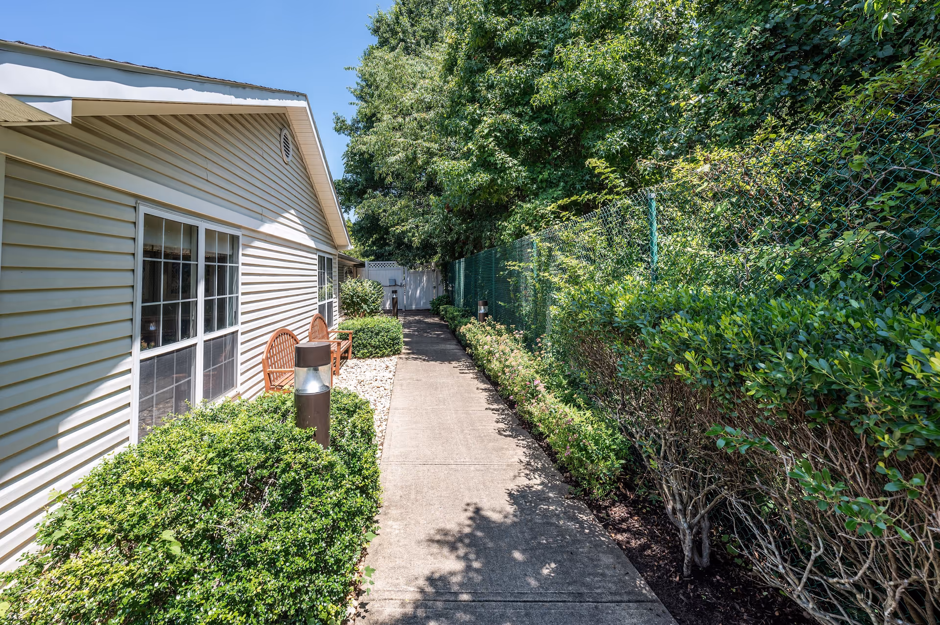 A paved walkway runs alongside a beige building with white-framed windows. On the left side of the walkway, there are green bushes and two wooden chairs. On the right side, there is a green chain-link fence with dense greenery and trees behind it. The sky is clear and blue.