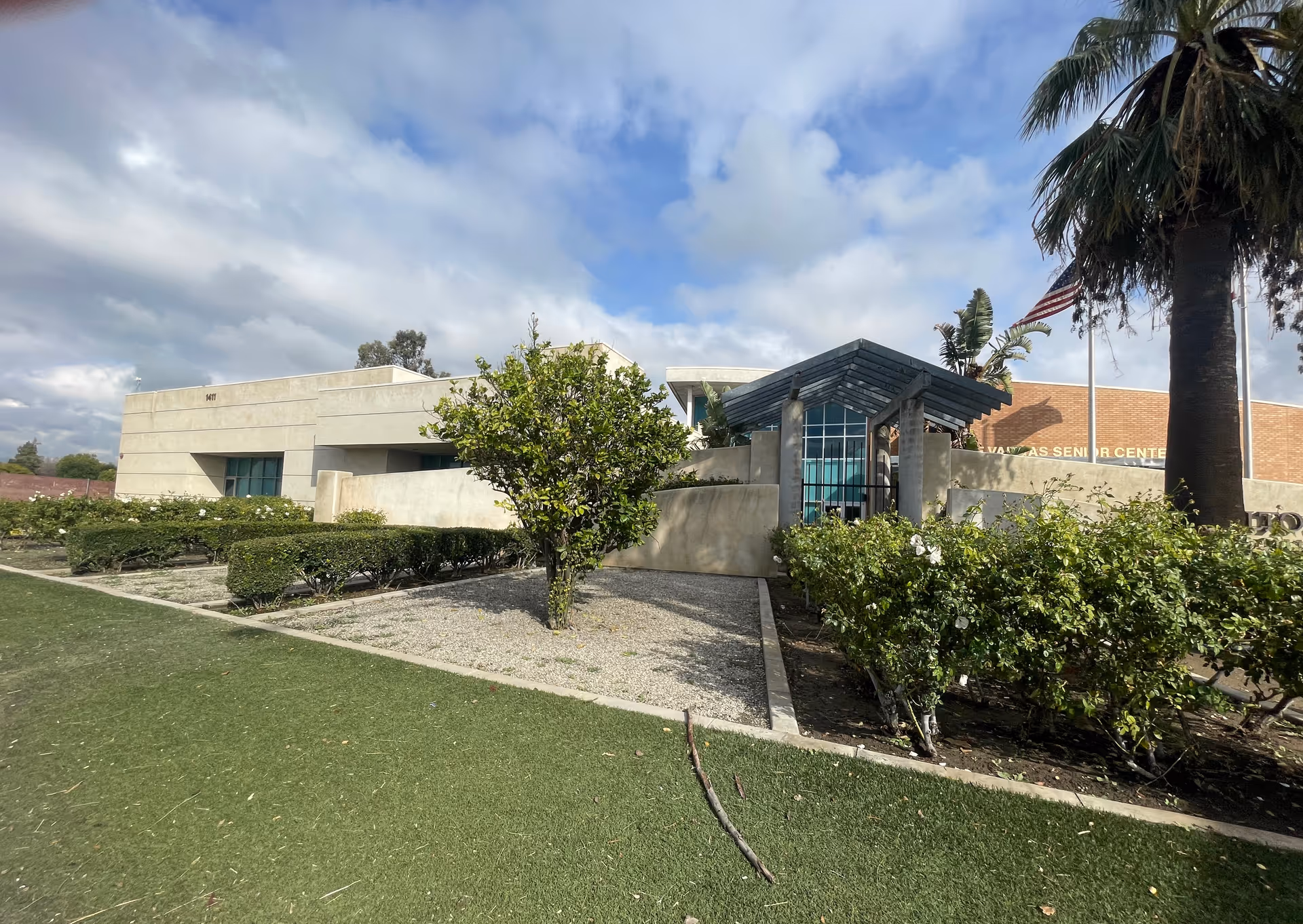 Exterior view of a modern building with beige walls and large windows, surrounded by green bushes, a palm tree, and a lawn under a partly cloudy sky. An American flag is visible near the entrance.
