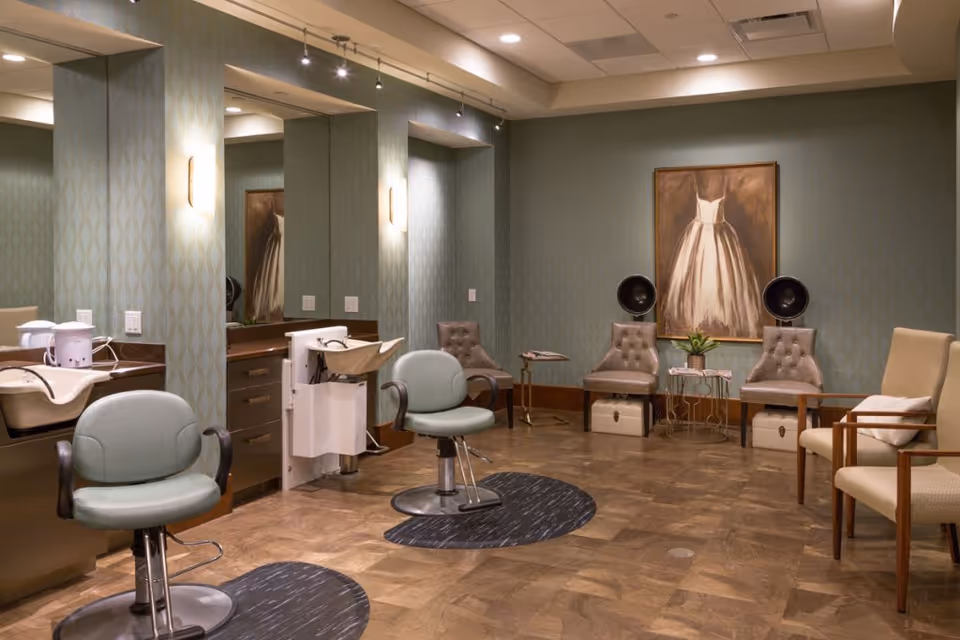 Interior of a salon area with two green salon chairs in front of mirrors and sinks on the left side. On the right side, there are several upholstered chairs, two vintage hair dryers, a small table with a plant, and a large framed painting of a white dress on the wall. The room has patterned wallpaper and wood flooring.