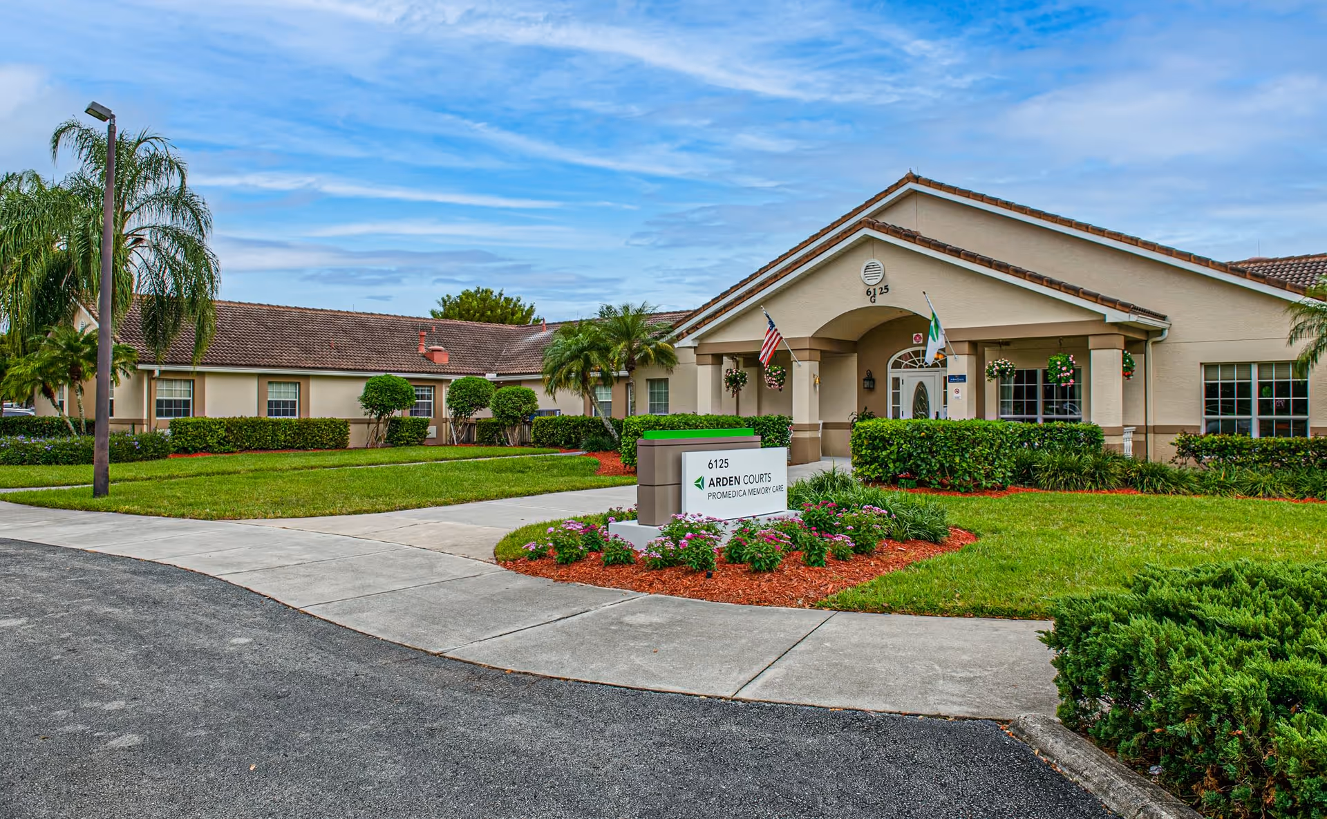 Exterior view of a single-story senior living facility building with a well-maintained lawn, palm trees, and a sign that reads '6125 Arden Courts ProMedica Memory Care'. The building has a beige facade with a tiled roof and an entrance with two flags and hanging flower baskets.