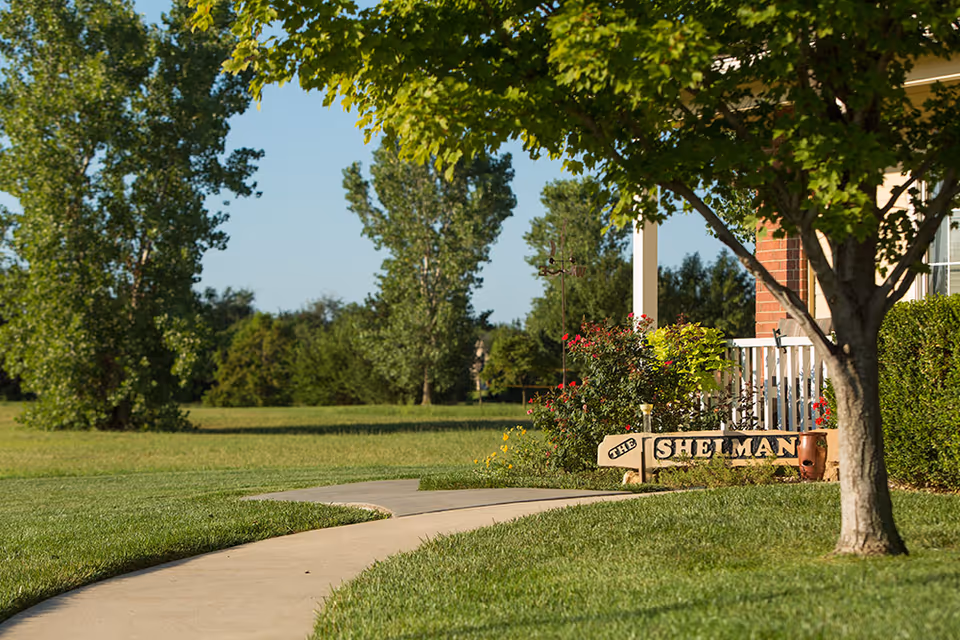 A curved concrete pathway leads through a well-maintained grassy area with trees and bushes. To the right, there is a partial view of a brick building with a white porch railing and a wooden sign that reads 'THE SHELMAN' surrounded by flowers and greenery.