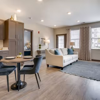 A bright and modern living room area with a white sofa adorned with blue and gray pillows, a gray rug on light wood flooring, a round dining table with two black chairs, and large windows with beige curtains letting in natural light. The room also features recessed ceiling lights, a small side table with a lamp, and a partial view of a kitchen area with brown cabinets.