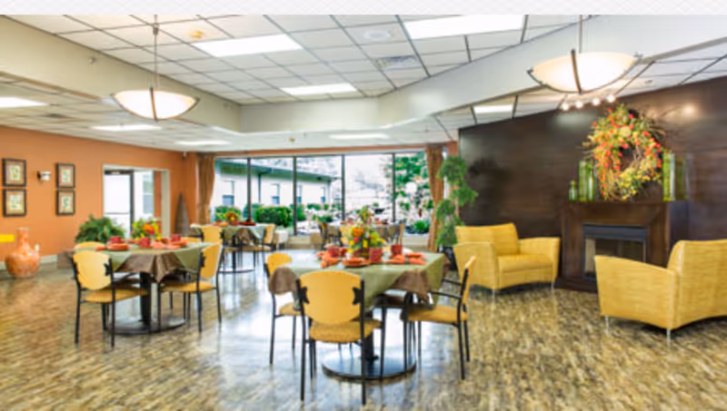 A bright and spacious dining area in a senior living facility with several round tables covered with green and brown tablecloths, each set with red napkins and floral centerpieces. The room features large windows letting in natural light, yellow cushioned chairs, a seating area with yellow armchairs near a dark wood fireplace decorated with a large floral wreath, and plants placed around the room.