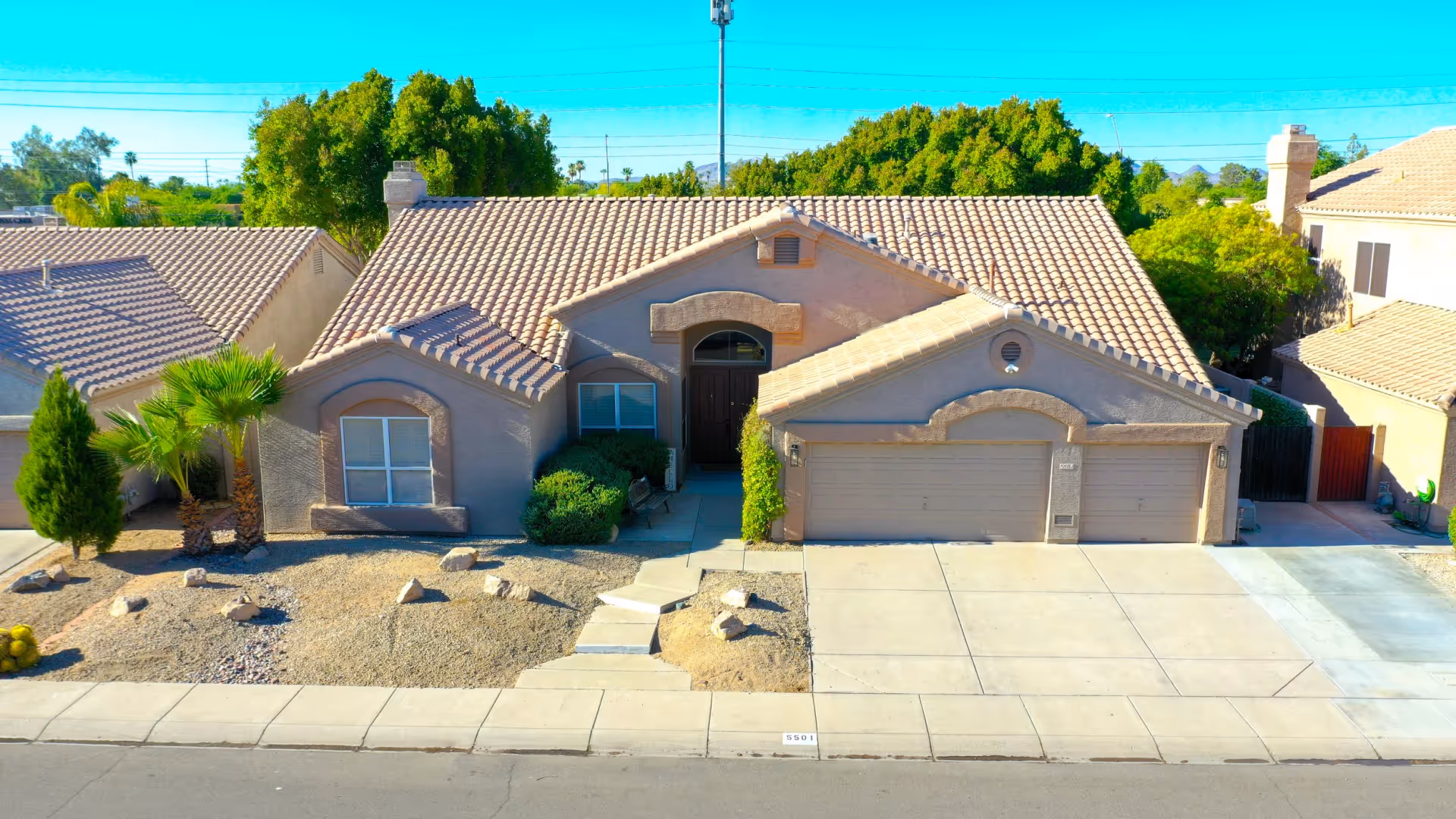 Single-story stucco house with a tiled roof, three-car garage, driveway and desert landscaping in front.