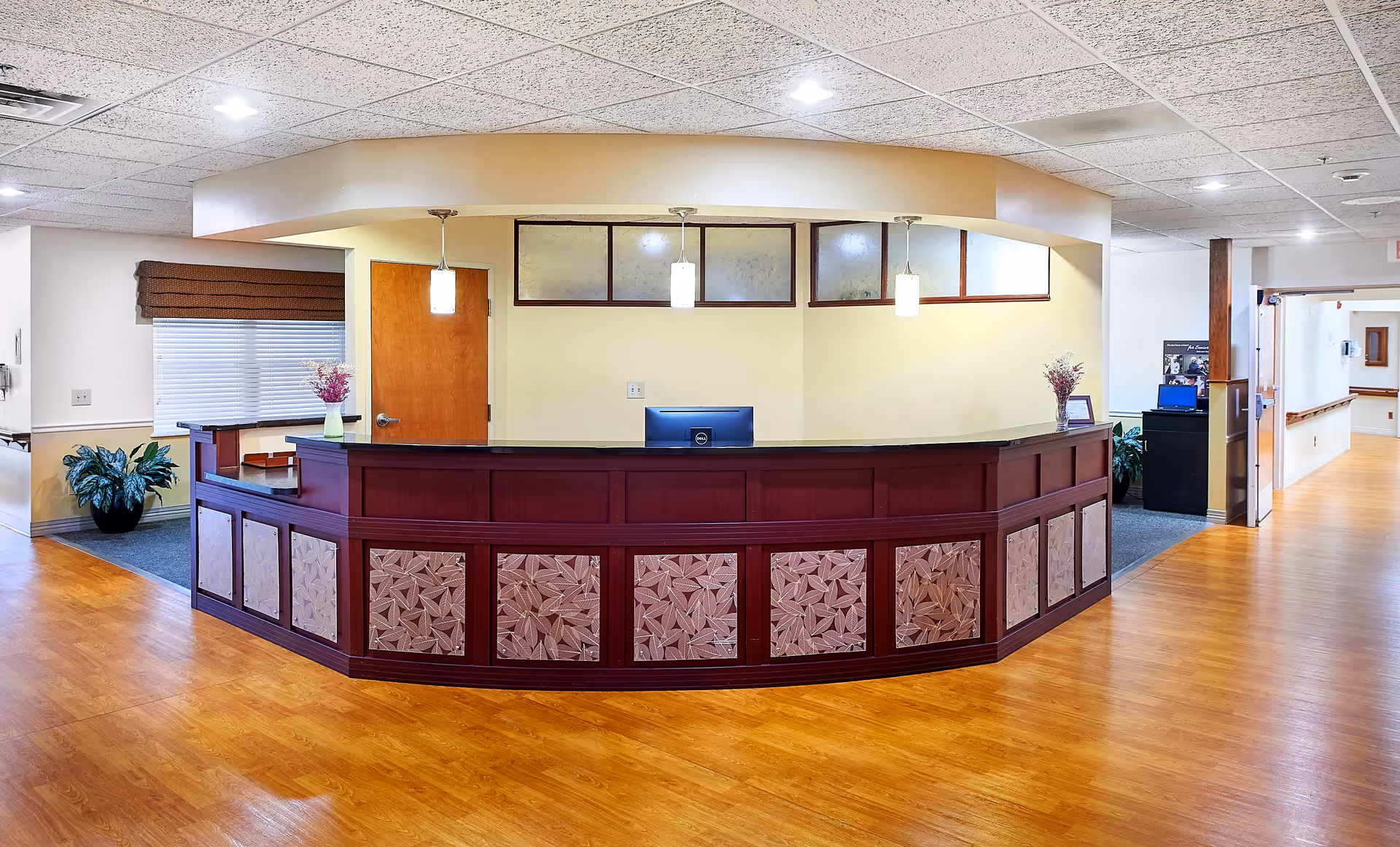 Reception desk area in a senior living facility with a wooden counter featuring decorative panels, three pendant lights hanging above, a computer monitor on the desk, potted plants on either side, and a hallway extending to the right.
