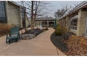 Outdoor paved walkway between two buildings with leafless trees and shrubs on either side. There is a green bench on the left side and a covered patio area with chairs and a closed umbrella in the background under an overhang.
