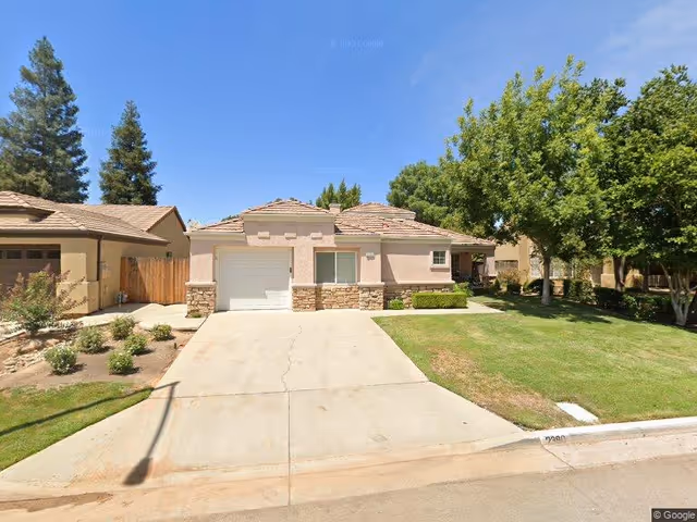 Single-story residential building with a beige exterior, stone accents, a garage, and a driveway. The house is surrounded by a well-maintained lawn and trees under a clear blue sky.