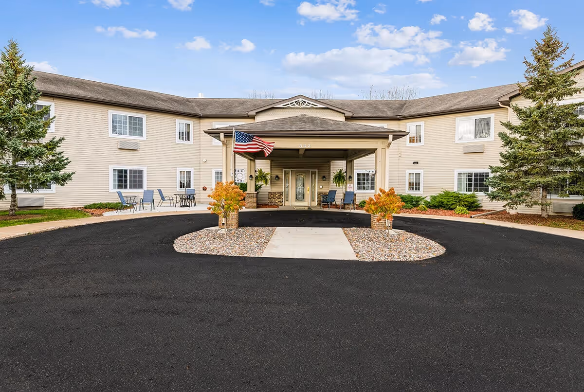 Front exterior view of Oak Gardens Place, a two-story beige building with a covered entrance, American flag, outdoor seating, and landscaped area with small trees and shrubs under a partly cloudy sky.