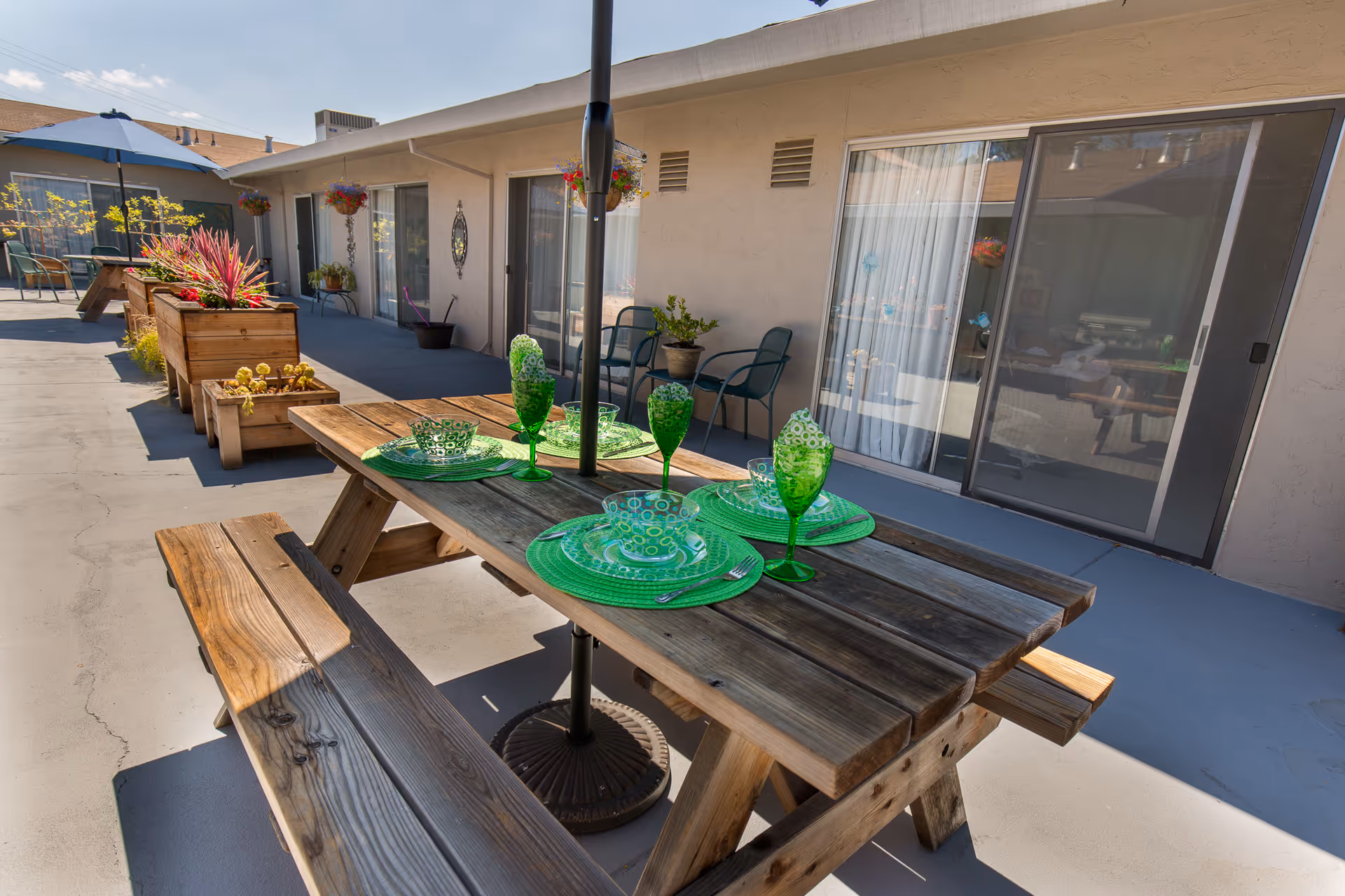 Outdoor patio area at Windsor Vallejo Care Center with wooden picnic tables set with green plates, bowls, and glasses. There are potted plants and hanging flower baskets along the building wall with sliding glass doors leading inside.
