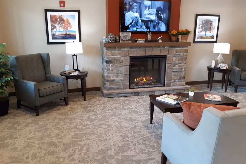 A cozy living room area in Country Meadow Place featuring a stone fireplace with a TV mounted above it showing a black and white movie. There are two armchairs with side tables and lamps on either side of the fireplace, a coffee table with magazines, and a light blue armchair with an orange pillow in the foreground. The walls have framed artwork and there is a patterned carpet on the floor.