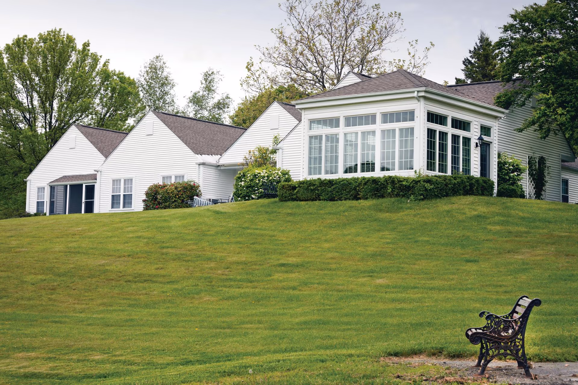 White single-story senior living building with a large sunroom on a grassy hill and a bench in the foreground.