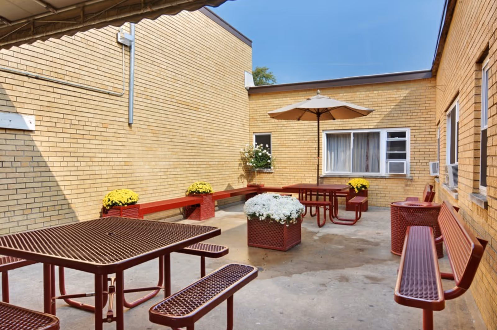 Outdoor courtyard area with red metal picnic tables and benches, yellow brick walls surrounding the space, potted flowers including white and yellow blooms, and a large beige patio umbrella providing shade over one of the tables.